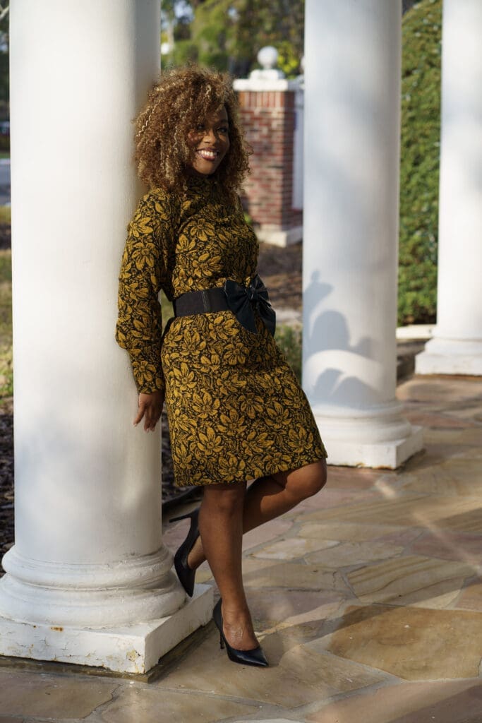 Petite sewist leaning against a white column wearing a mustard floral matching set Simplicity 8982 with a black waist belt.