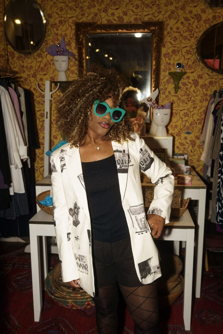 Close up of a Desigual outfit idea featuring a graphic newsprint blazer and a pop of color with teal cat-eye sunglasses.