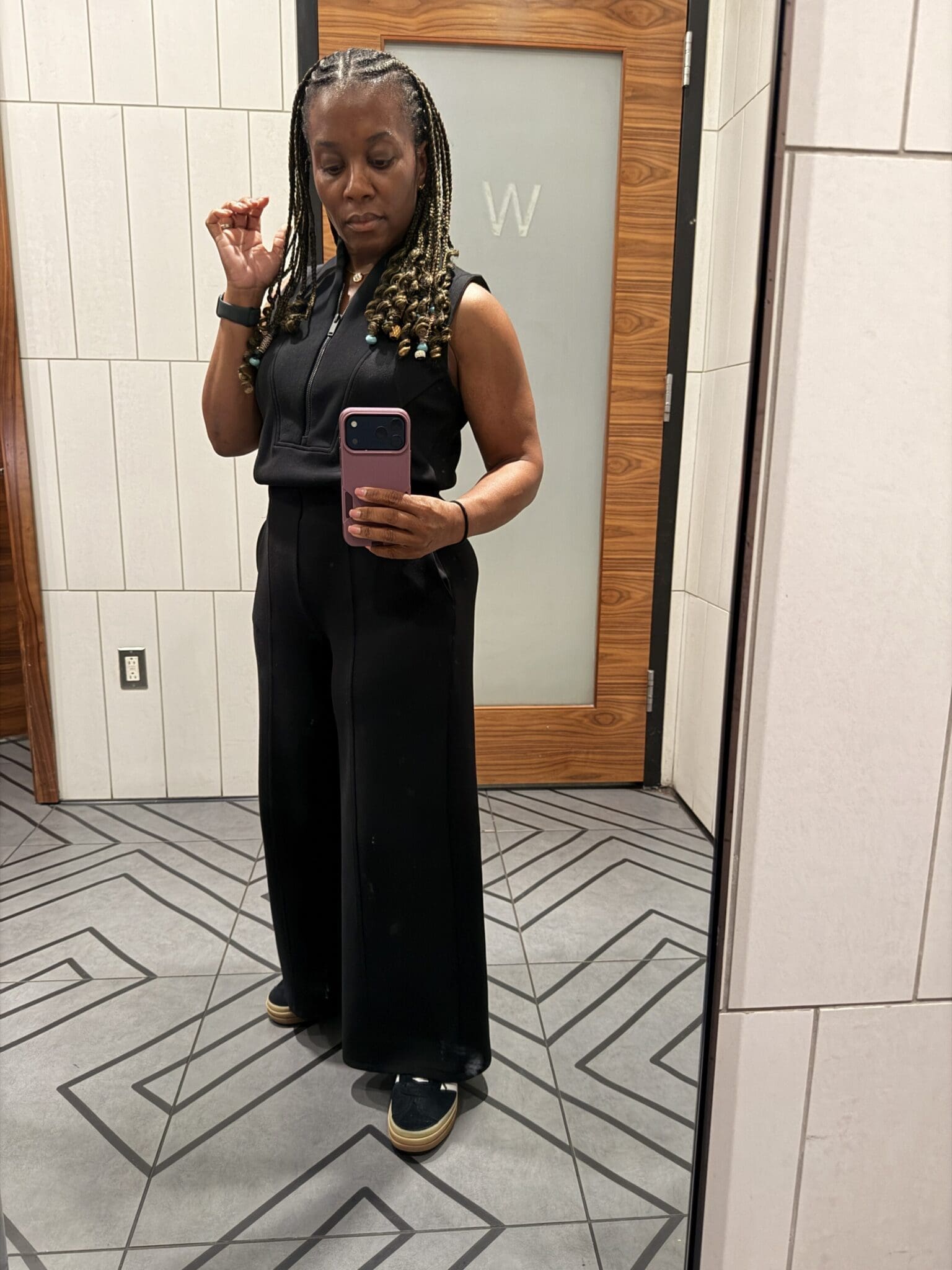 Woman posing in a modern airport lounge wearing a comfortable black sleeveless Spanx AirEssentials travel jumpsuit.